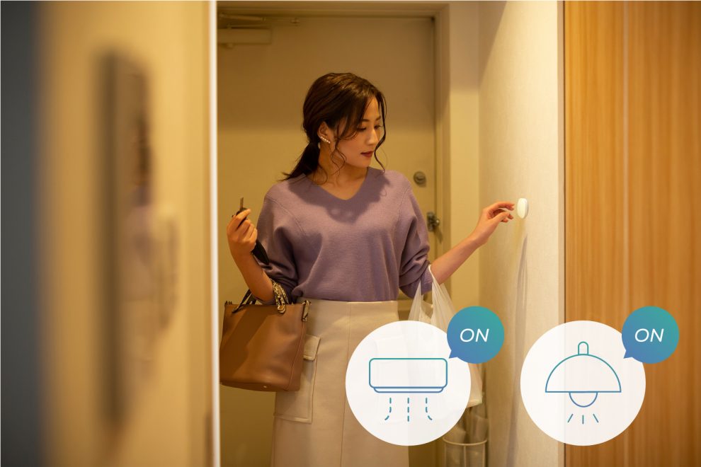 帰宅した女性が玄関の壁に設置したスマートボタンを押すイメージ